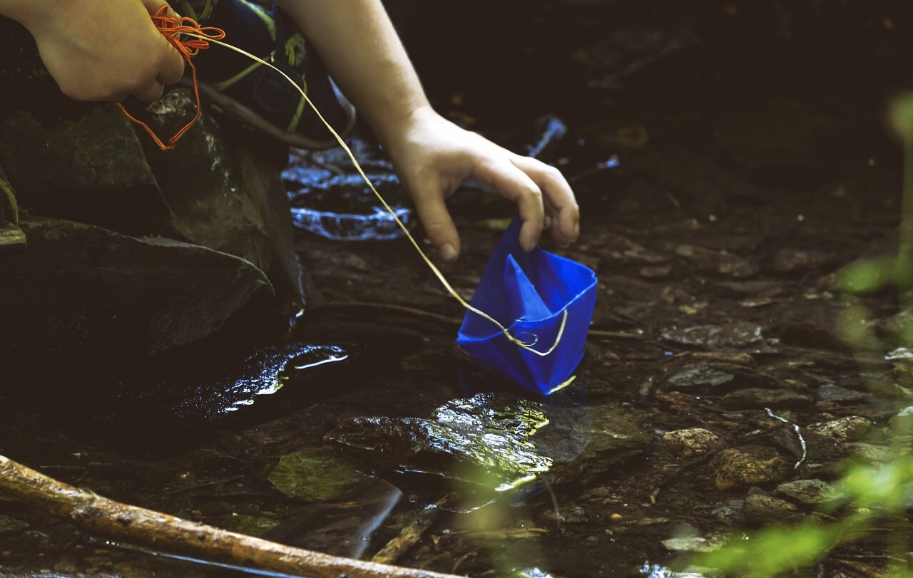 Child plays with blue paper boat by a stream