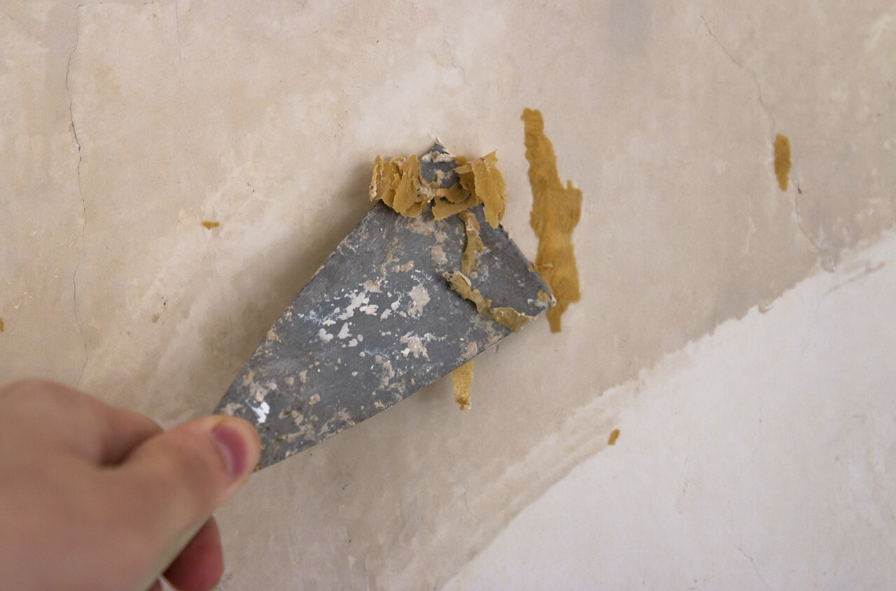 Hands removing old wallpaper with the help of a spatula during the repair in the room.